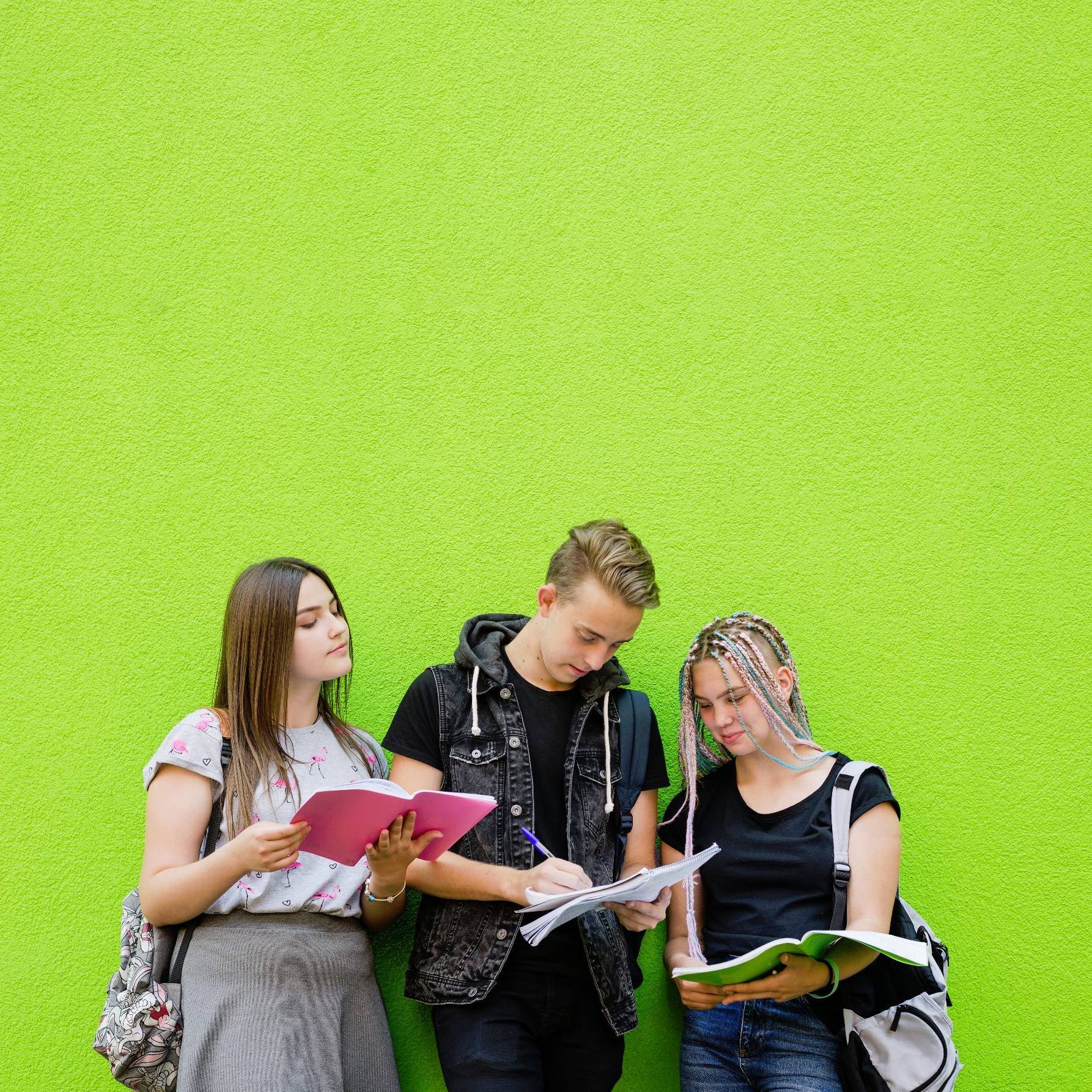 Drei Schüler*innen stehen vor einer grünen Wand und schauen gemeinsam in ihre Hefte